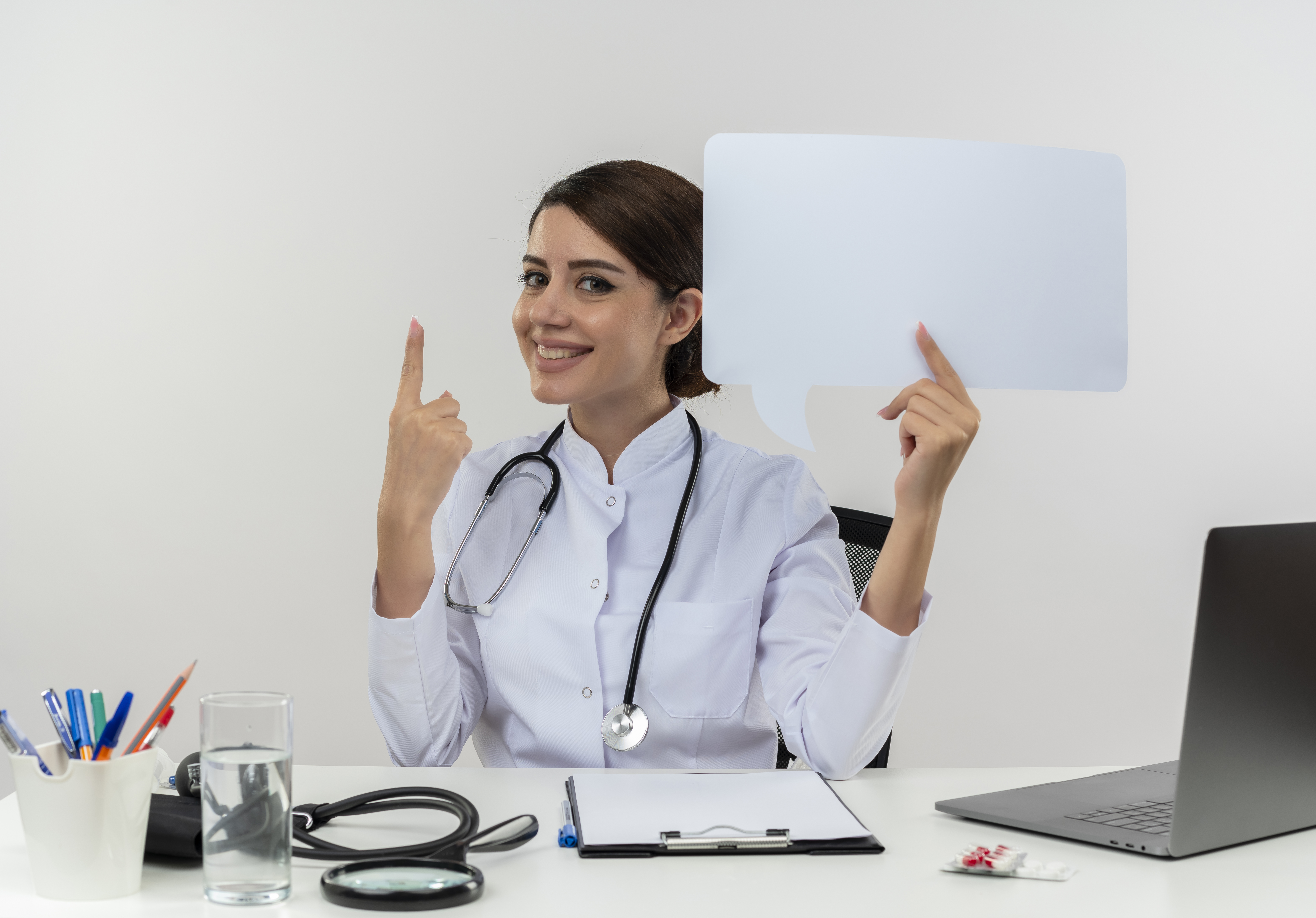 smiling young female doctor wearing medical robe with stethoscope sitting at desk work on computer with medical tools holding chat bubble poiunts to up on isolated white background with copy space